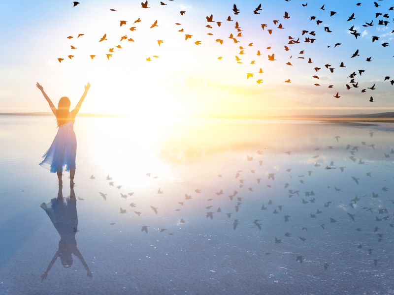 Angst vor der Angst überwinden: Eine Frau steht mit erhobenen Armen am Wasser bei Sonnenaufgang und blickt auf einen Vogelschwarm als Symbol für Freiheit und psychische Gesundheit in der Therapie.