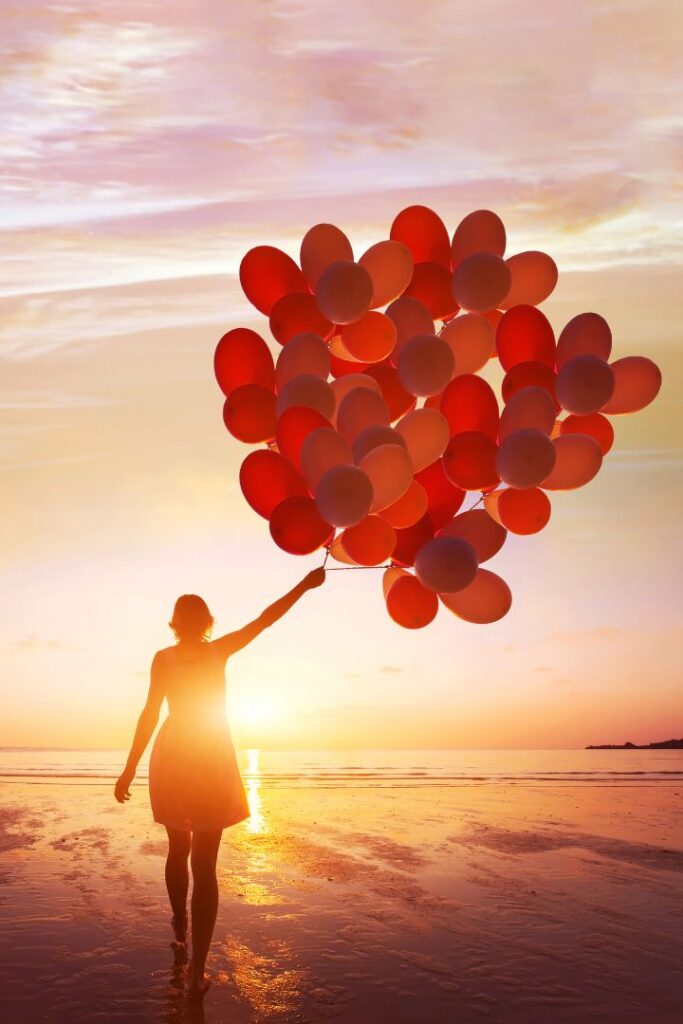 Frau am Strand hält bei Sonnenuntergang ein Bündel roter Luftballons als Symbol für emotionale Freiheit durch Selbsthypnose lernen in Rudolstadt.