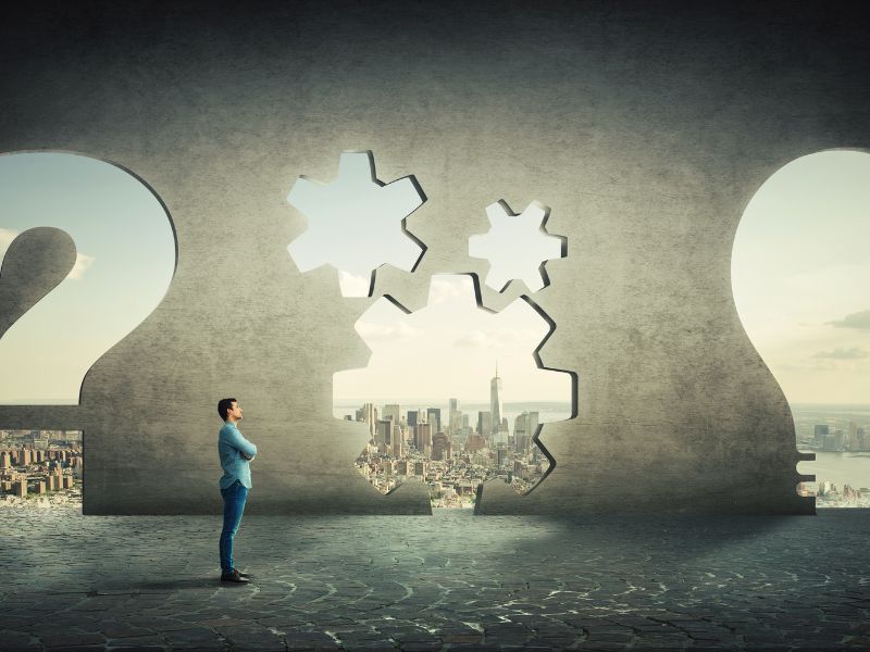 Ein Mann steht vor einer Betonwand mit Aussparungen in Form von Zahnrädern und blickt auf eine Stadt, als Metapher für die Suche nach der Lösung durch Ursachenorientierte Hypnose Thüringen.